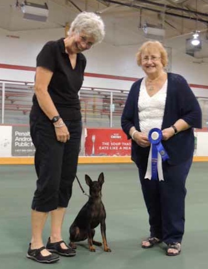 Two women stand with a small dog between them; one holds a blue ribbon.