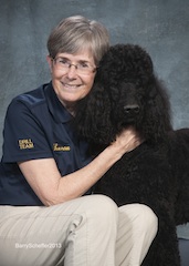 Person wearing a navy polo shirt smiling and holding a large black poodle against a gray backdrop.
