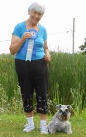 A woman holds a blue ribbon next to a small schnauzer dog in a grassy area.