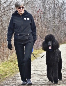 A woman wearing sunglasses and a black jacket walks a black poodle on a path through a leafless forest.