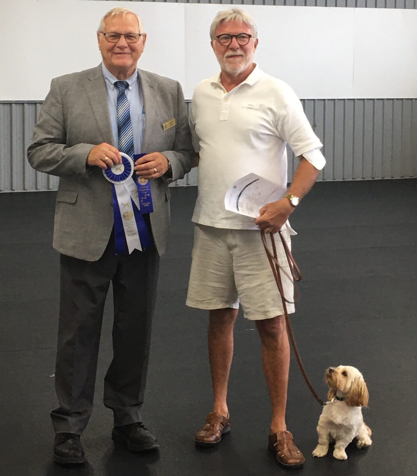 Two men standing together indoors; one holds a blue ribbon and the other holds a small dog on a leash.