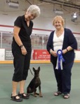 Two women stand beside a small dog on an indoor field. One woman holds a blue ribbon.